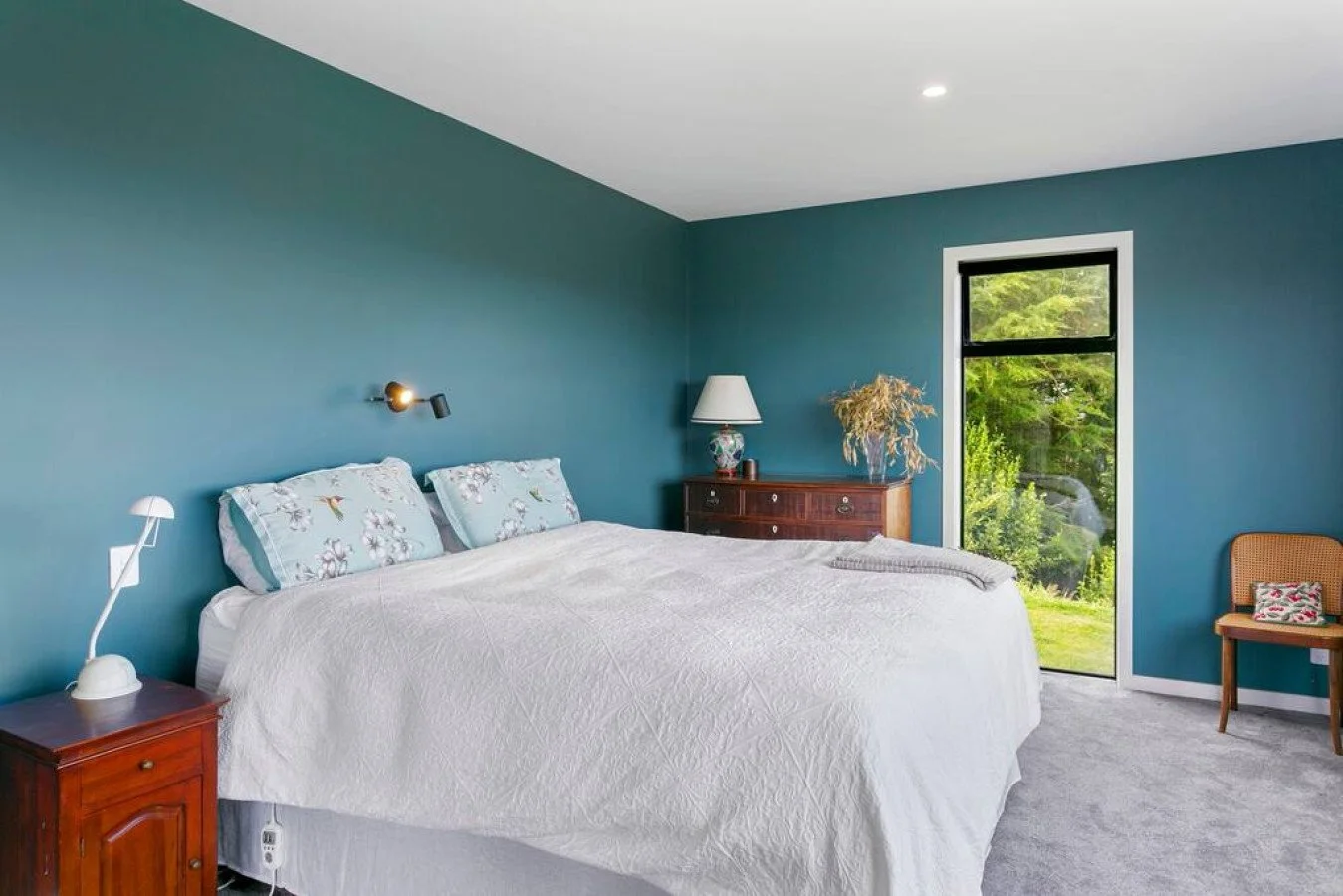 A bedroom with teal walls, a white bed with floral pillows, a wooden dresser with a lamp and plant, a window with green outdoor scenery, and a wooden chair.