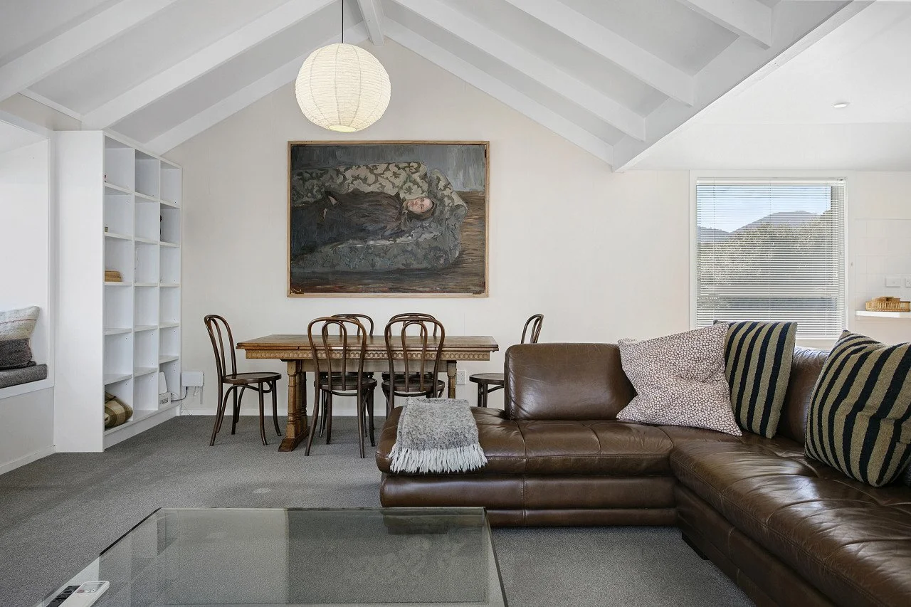 Living room with a brown leather sectional sofa including pillows, a glass coffee table, a dining table with six chairs, a large framed portrait painting on the wall, a white bookshelf, a paper lantern ceiling light, and a window with blinds showing 