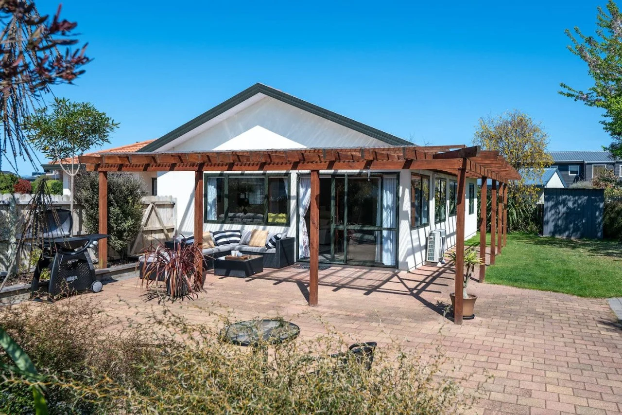 A backyard patio with a white house, a wooden pergola, patio furniture, a barbecue grill, and surrounding trees under a clear blue sky.