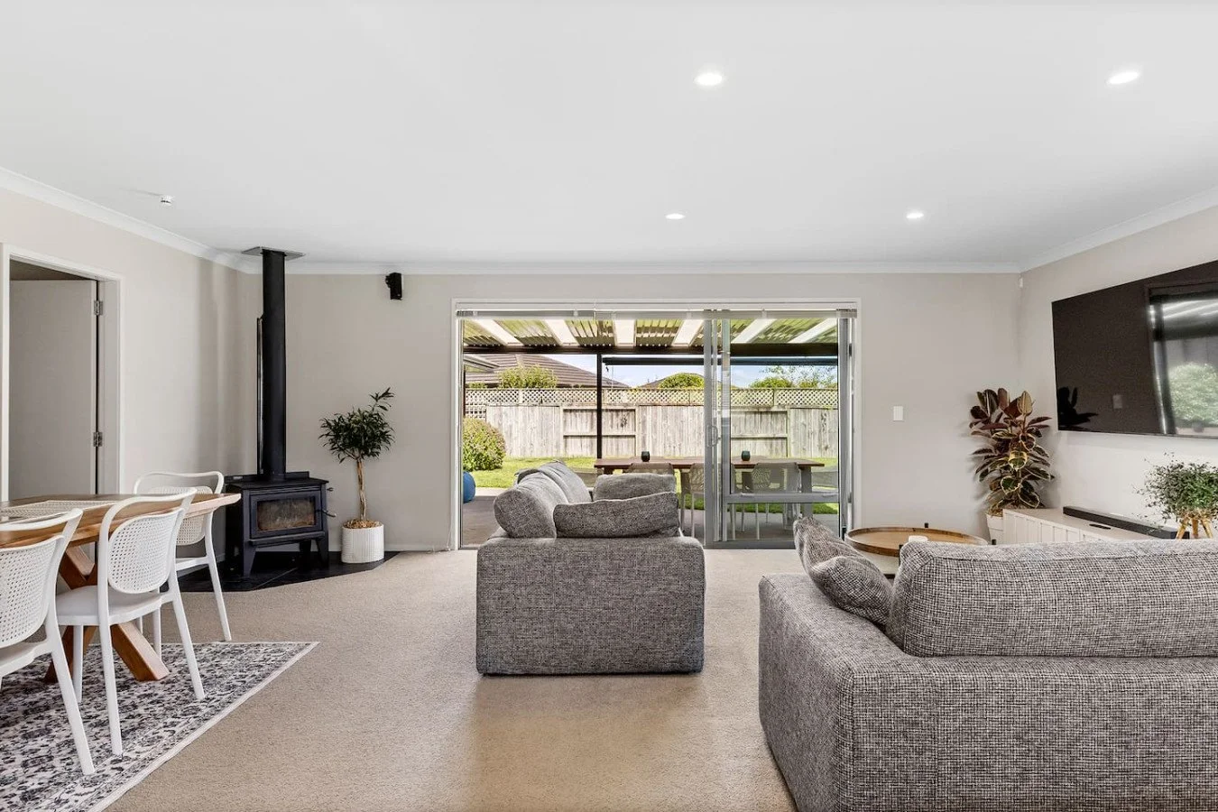 Living room with grey sofas, a TV on the wall, and an open sliding door leading to a backyard with outdoor furniture and a fence.