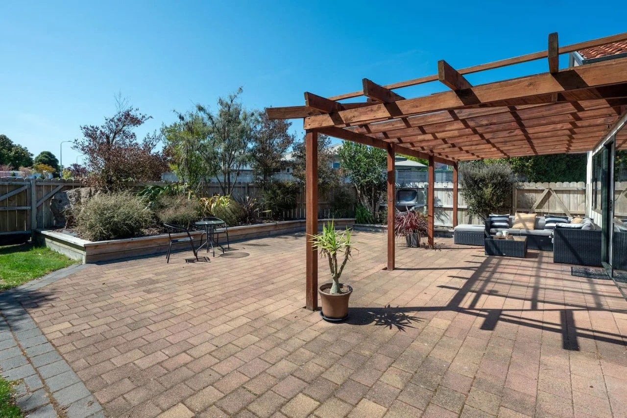 Spacious backyard patio with brick pavers, a wooden pergola, outdoor seating, potted plants, and a garden bed with bushes and trees under a clear blue sky.