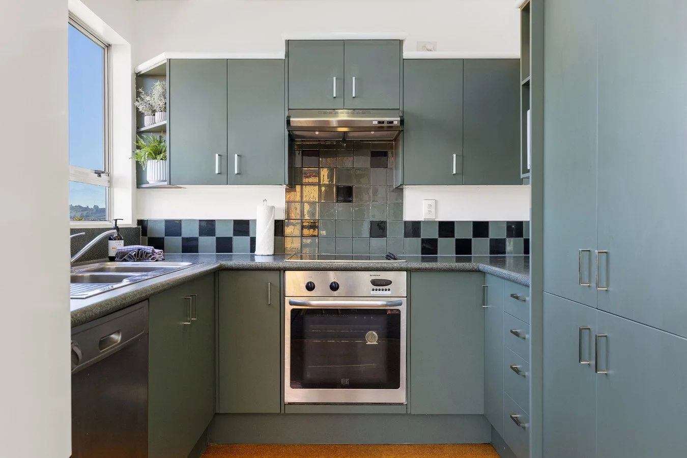 A compact kitchen with green cabinets, a tiled backsplash, a stainless steel oven, a stainless steel exhaust hood, a window with a view, and a countertop with a paper towel roll and cleaning supplies.