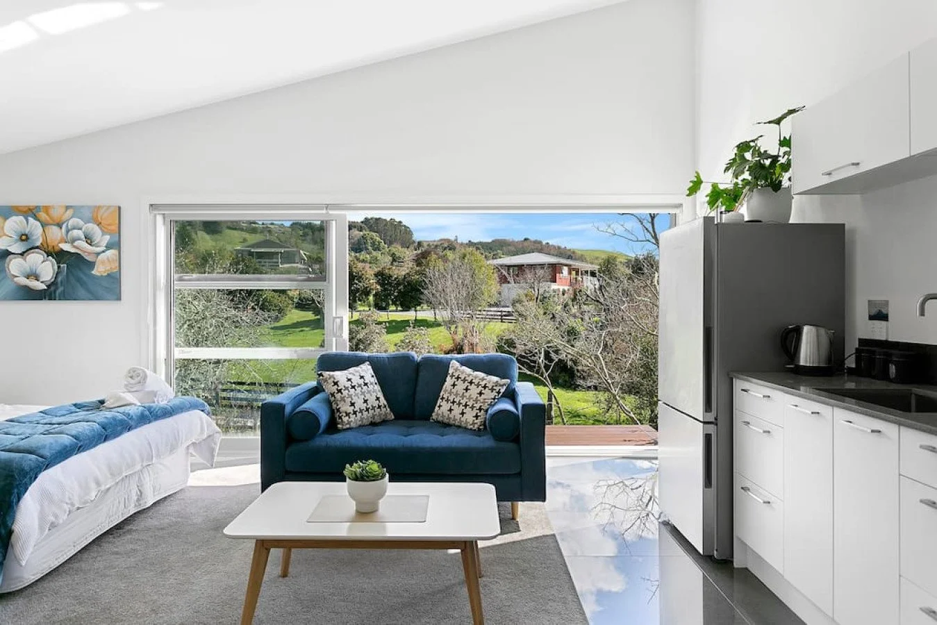 Living room with a large window overlooking a green landscape, a blue couch with patterned pillows, a white coffee table with a small plant, a bed with white linens and a blue blanket, artwork on the wall, and a kitchen area with white cabinets and a
