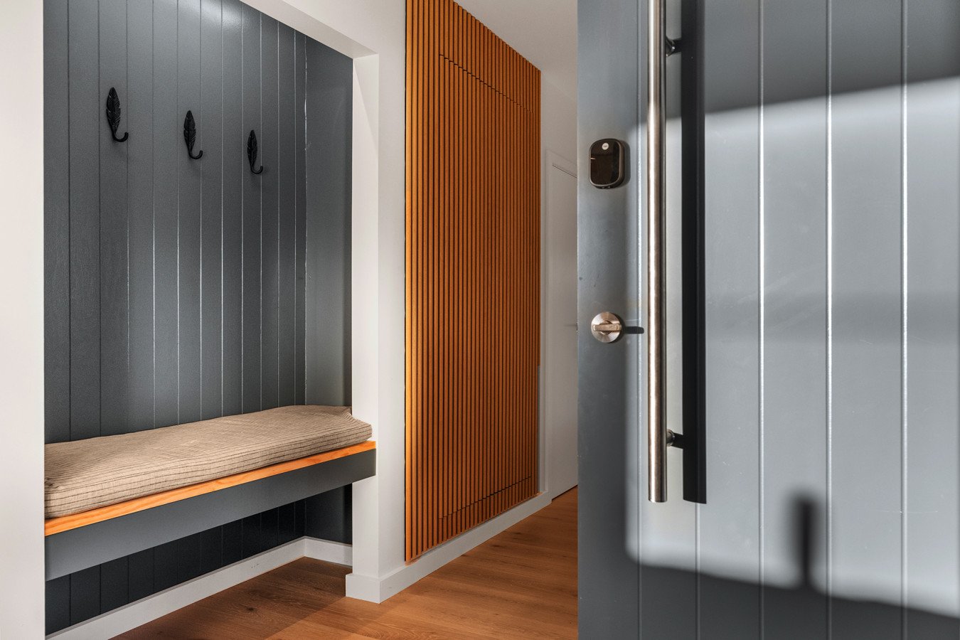 View of a modern entryway with a wooden bench and coat hooks on a dark gray wall, a wooden slatted panel, and a gray door with a metal handle and security lock.