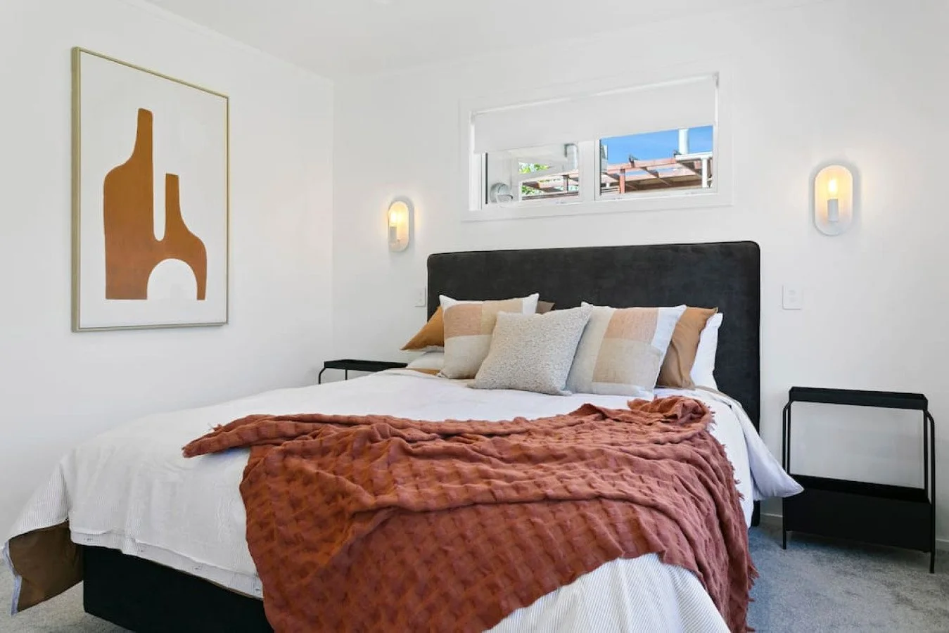 A neatly made bed with a black headboard, multiple beige and white pillows, and a rust-colored textured throw blanket. The bedroom has white walls, wall-mounted lights, a art piece on the wall, and a small black bedside table. There is a window above
