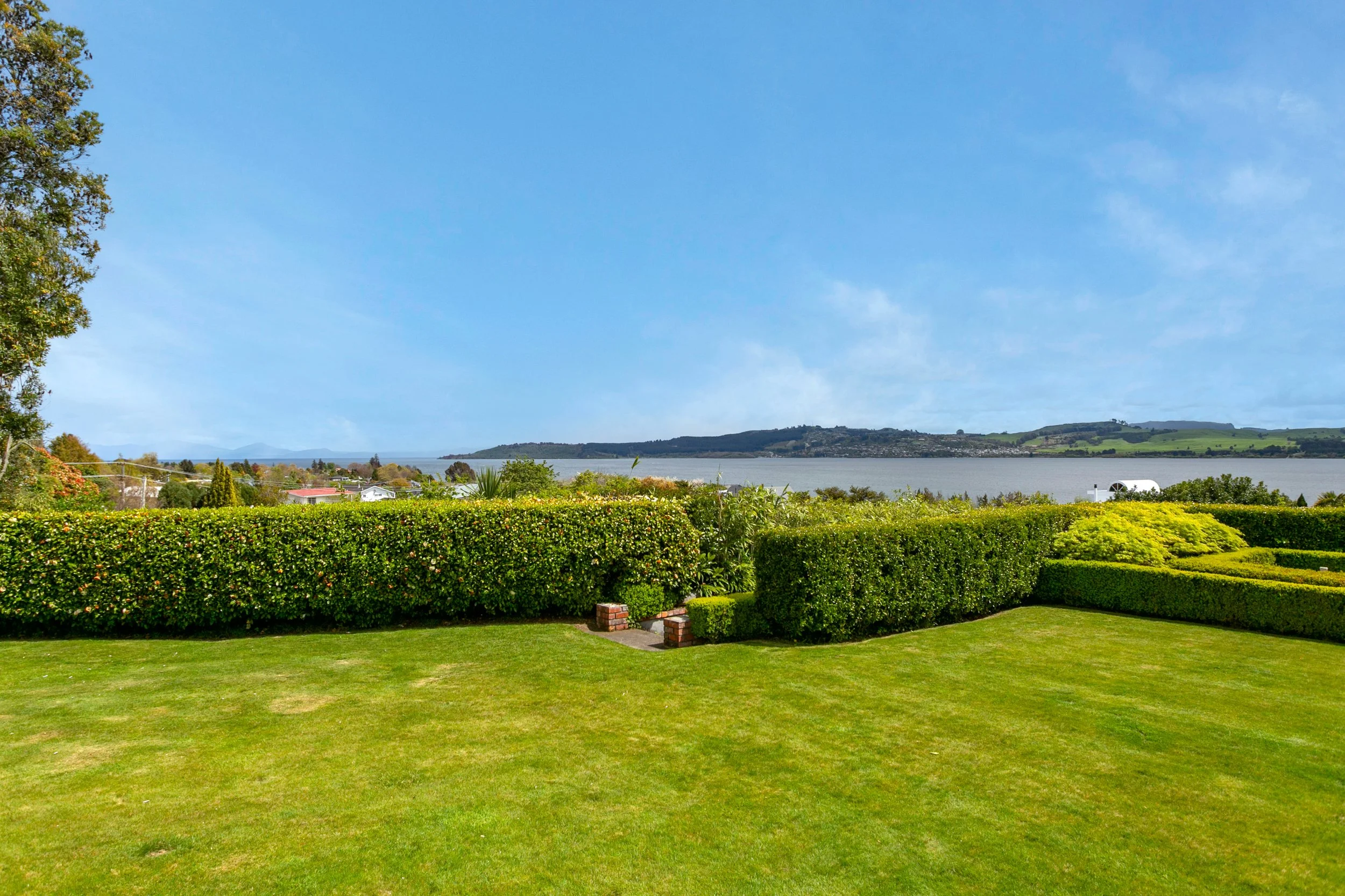 Landscape view of a manicured lawn, trimmed hedges, and a body of water in the distance under a partly cloudy sky.
