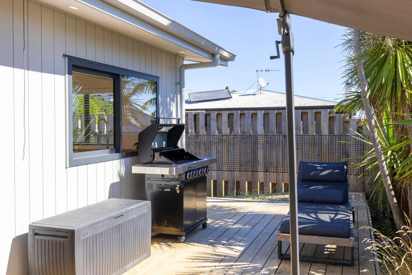 Outdoor patio area with a grill, lounge chair, storage bench, and umbrella pole, surrounded by wooden fence and plants, on a sunny day.