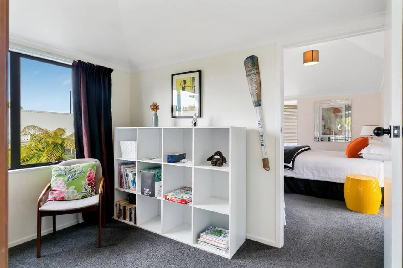 A cozy room with a window, black curtains, a white chair with a floral cushion, and a white bookshelf with books and decorative items. There is a doorway leading to a bedroom with a bed, orange pillow, yellow side table, and a mirror on the wall.
