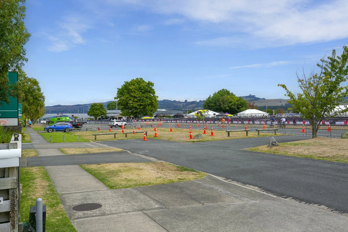 A parking lot near a waterfront with orange traffic cones, cars, and trees, with tents and hills in the background on a sunny day.
