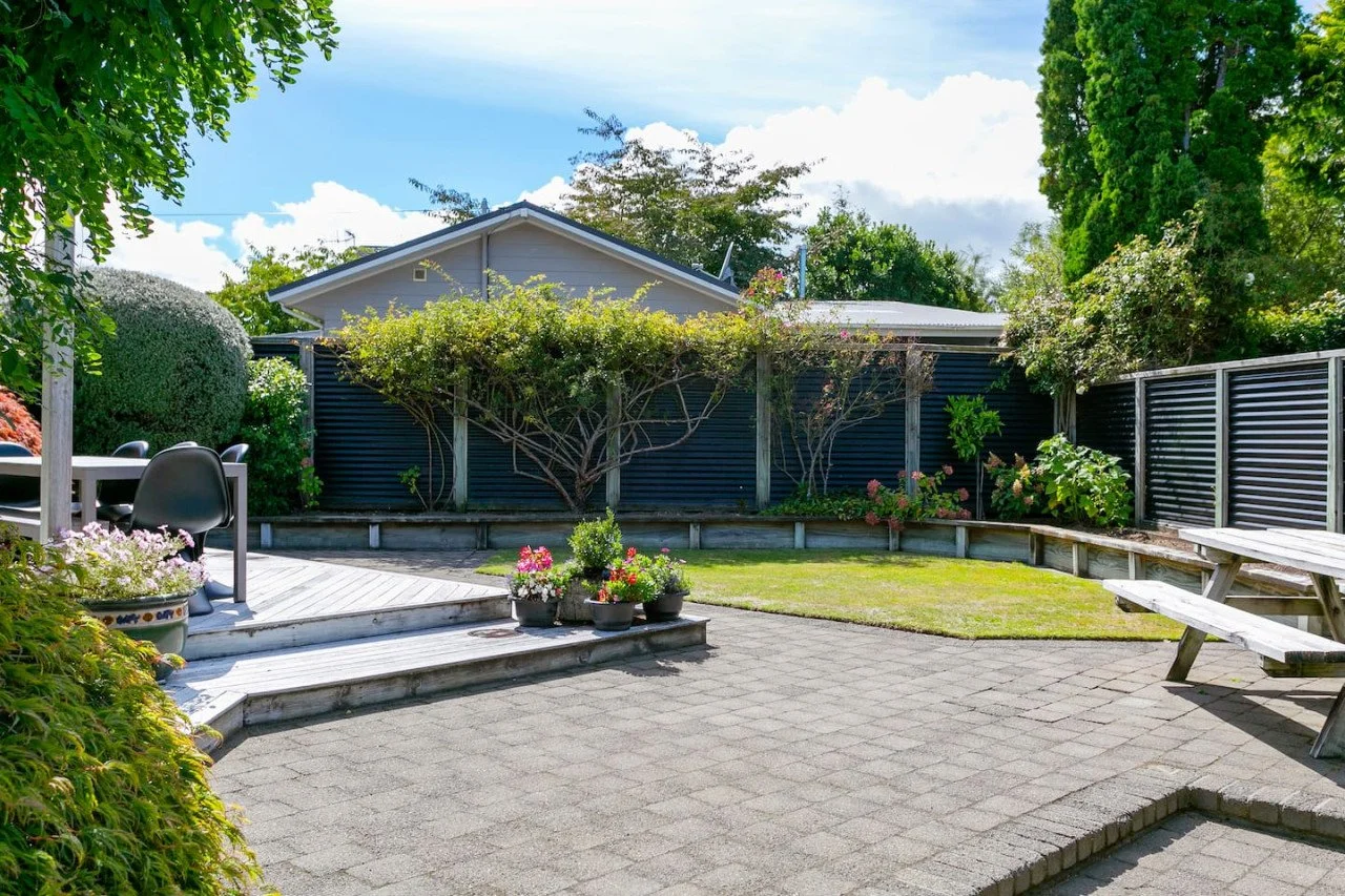 A backyard patio with a small grassy area, wooden terrace with black chairs, potted plants, a wooden picnic table with benches, and a tall fence with greenery and trees surrounding it.