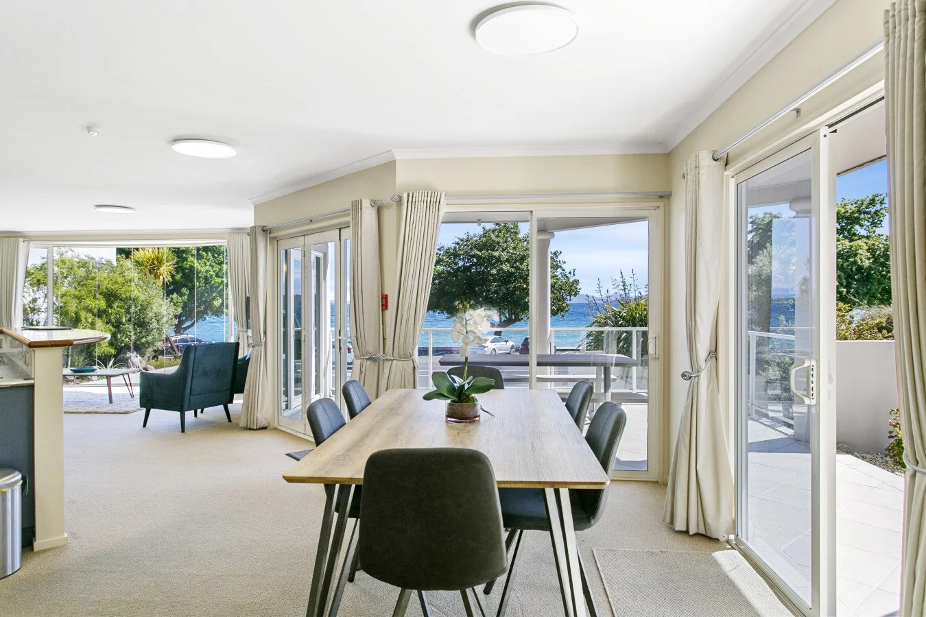 Living room with large glass windows and doors, ocean view, dining table with six chairs, potted white orchid, and outdoor patio.