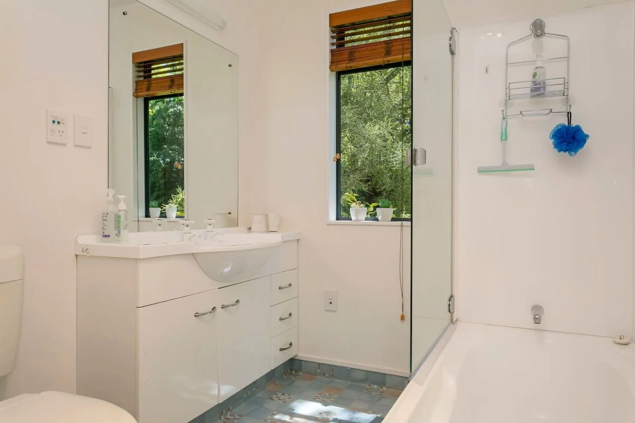Bright bathroom with a white vanity, mirror, window with wooden blinds, and a walk-in shower with a glass door. There are small potted plants on the window sill and a blue bath pouf hanging on a wall rack.