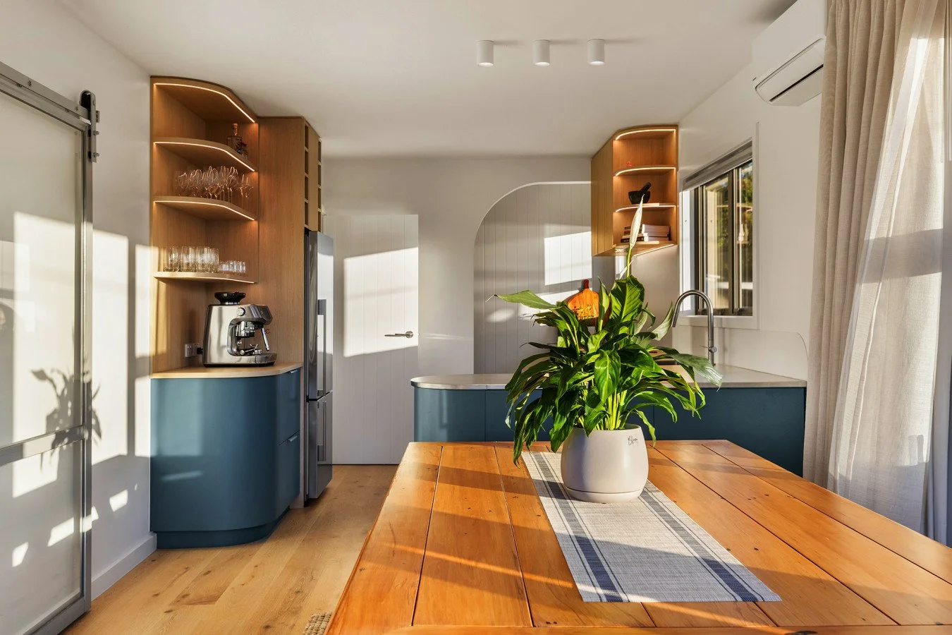 Bright kitchen with wooden dining table, potted plant, white curtains, and modern appliances including coffee maker and refrigerator.