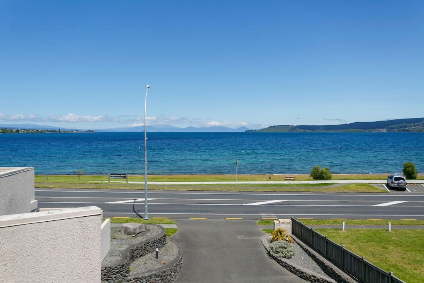 View of a large body of water, possibly a lake, with a grassy park area and a paved road in the foreground. There are a few small trees and bushes, a parked car, and a bench along the shoreline. Mountains are visible in the distance under a clear blu