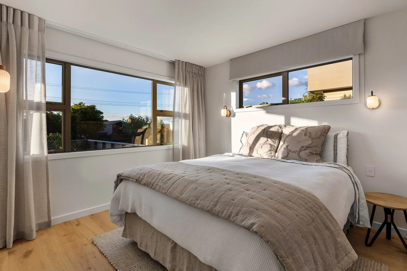 A bright modern bedroom with a large bed, beige and white bedding, and decorative pillows. Two wall-mounted lamps and large windows with sheer cream curtains let in natural light, revealing trees and a blue sky outside.