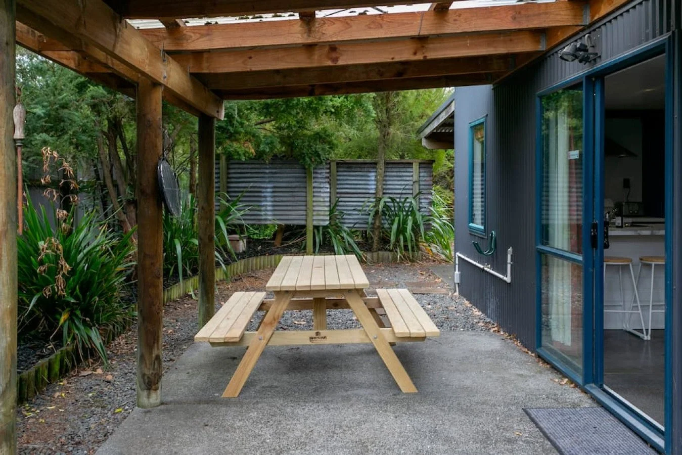 Wooden picnic table with attached benches under a wooden pergola, outdoors near a house with sliding glass doors, surrounded by plants and trees, with a metal fence in the background.