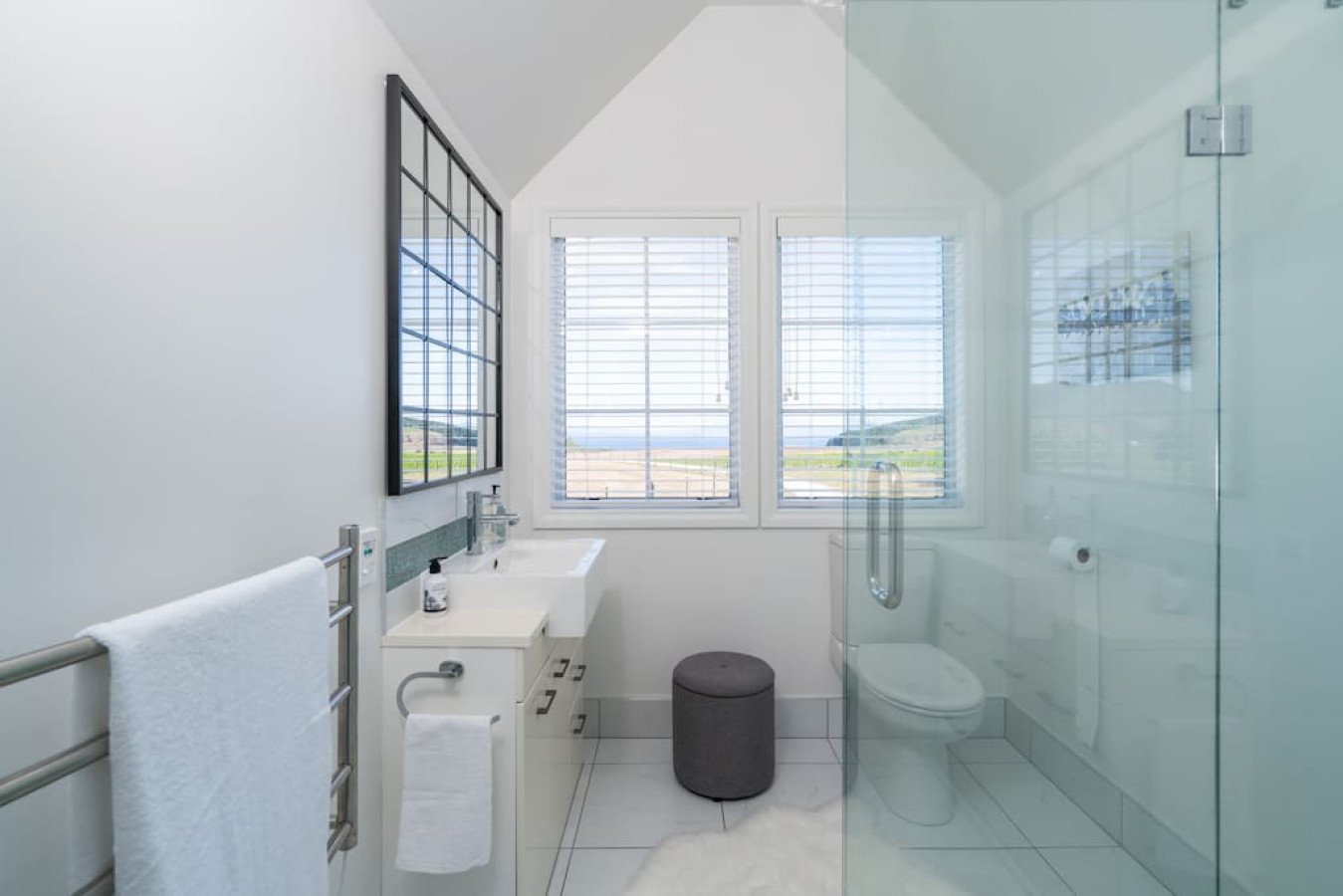 Bright bathroom with two windows, a white sink, a mirror, a towel rack, a small gray stool, and a toilet behind a glass shower enclosure.