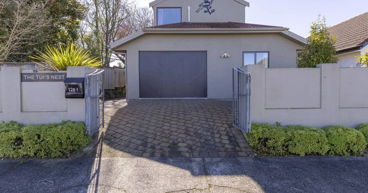 Gray house with a black garage door, metal gates, and a paved driveway, given a modern look with bushes and trees around