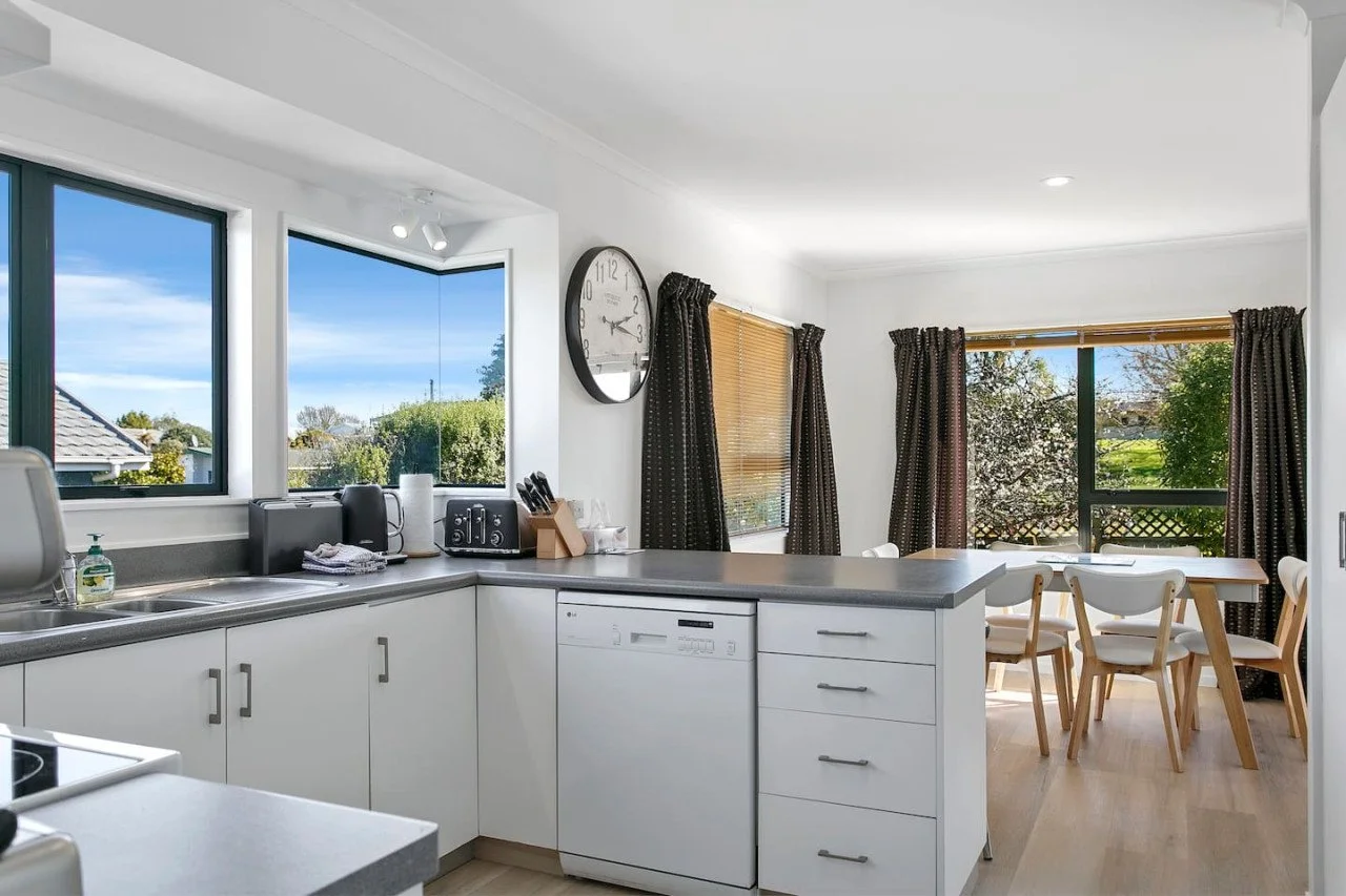 Kitchen with white cabinets, black countertops, large windows showing outdoor greenery, a round wall clock, a dining table with chairs, and various kitchen appliances and utensils.