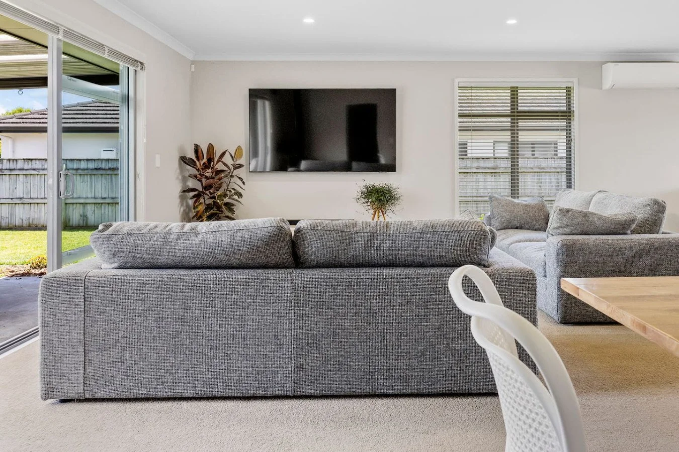 Living room with gray sofas, a wall-mounted flat-screen TV, large window with blinds, a sliding glass door leading to a backyard, indoor plants, and a wooden dining table with a white chair.