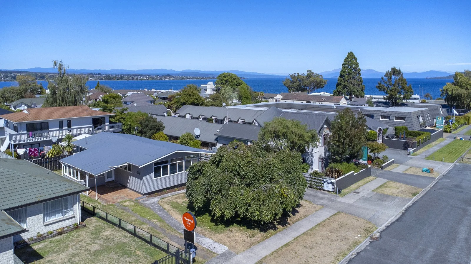 Residential neighborhood near a body of water with hills or mountains in the background, including single-family homes, trees, and a clear blue sky.