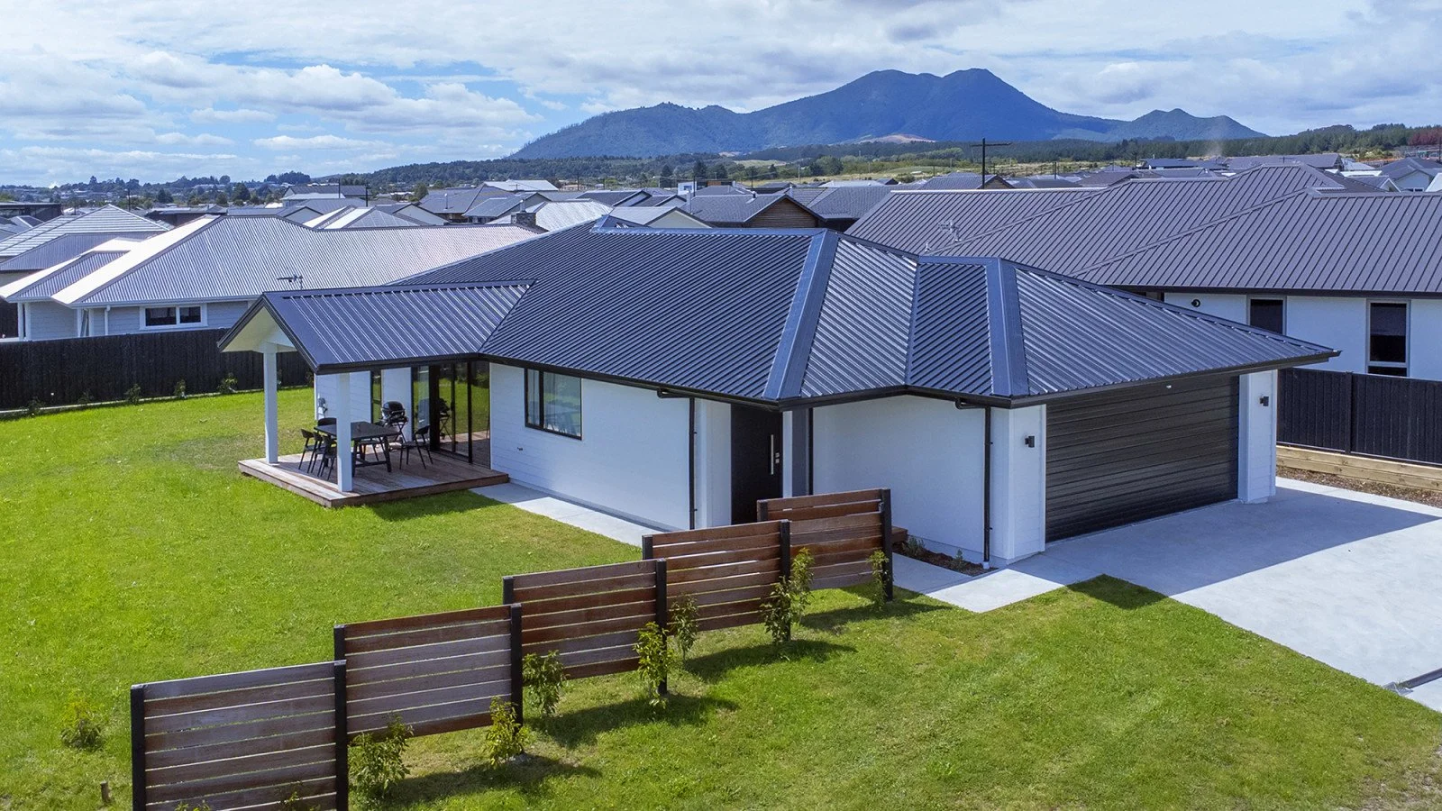 Modern house with metal roof and a backyard patio with outdoor furniture, green lawn, and wooden fence, with mountains in the background.