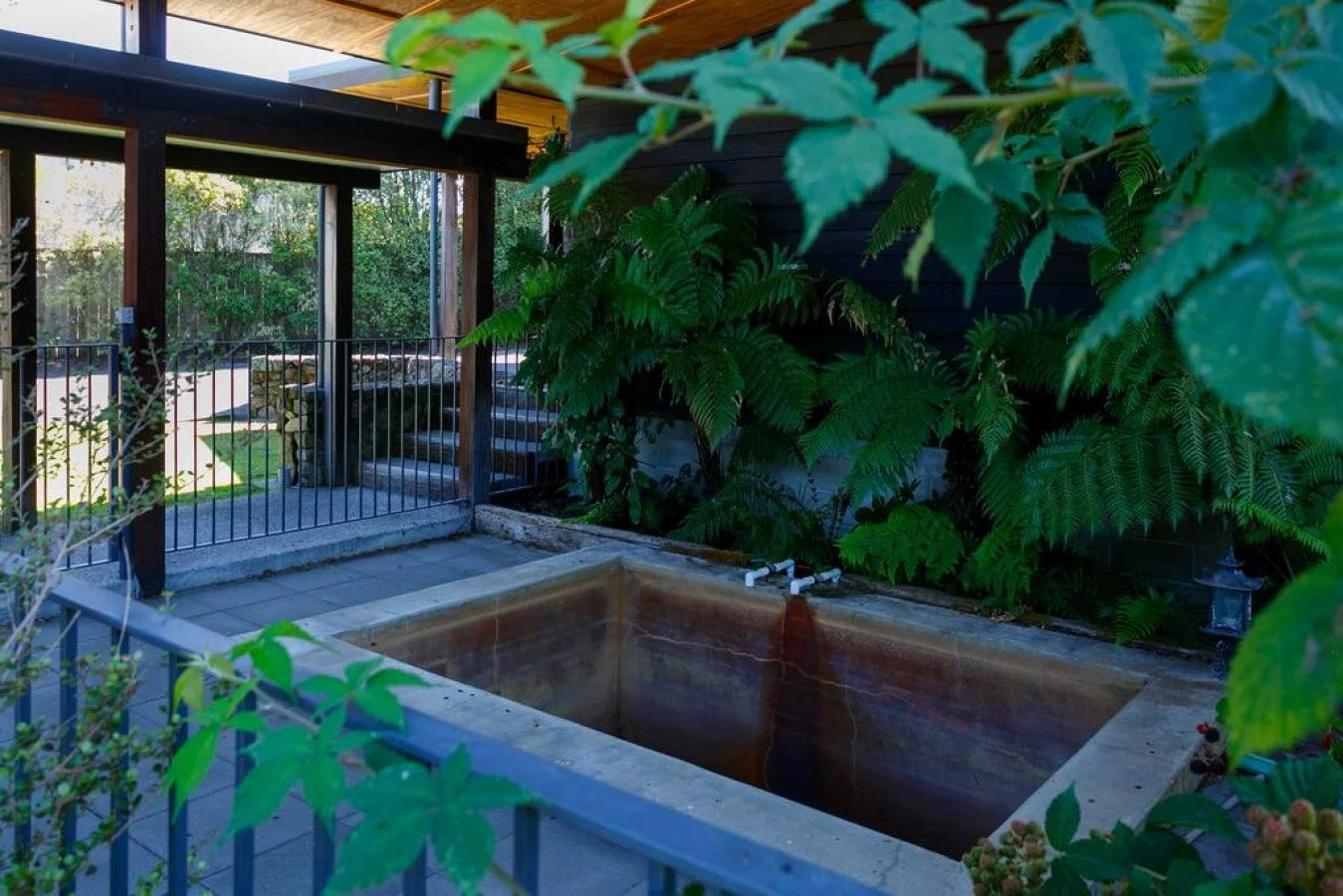 View of a small outdoor concrete hot tub or fountain, surrounded by a black metal fence, foliage, and an outdoor seating area in the background.
