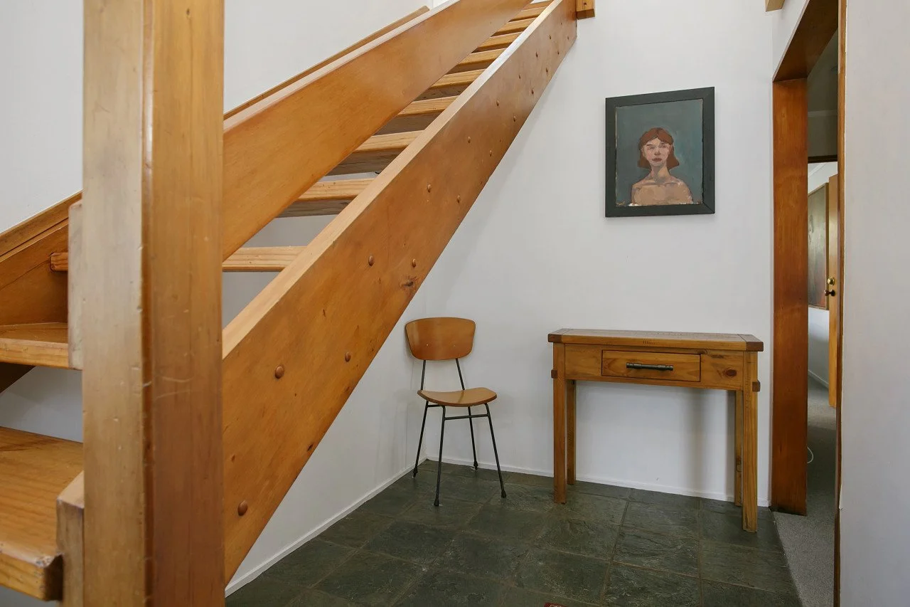 Interior view of a hall or stairwell with wooden staircase, a small wooden table, a chair, a portrait painting on the wall, and a doorway leading to another room.
