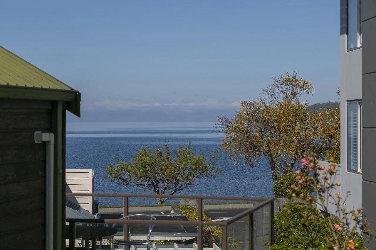 View of a calm blue lake or ocean through a balcony with trees, buildings on either side, and a clear blue sky