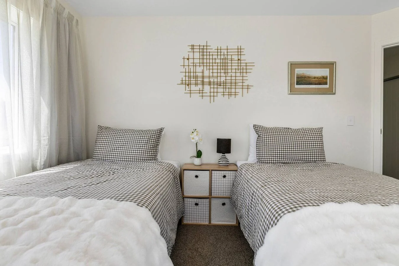Two beds with checkered bedding and pillows, a nightstand with a small potted plant and a striped lamp, framed artwork and wall decor above, sunlight through a curtain on the left.