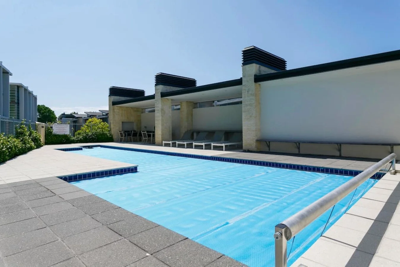 Empty rooftop swimming pool with lounge chairs and a shaded seating area, in sunny weather.