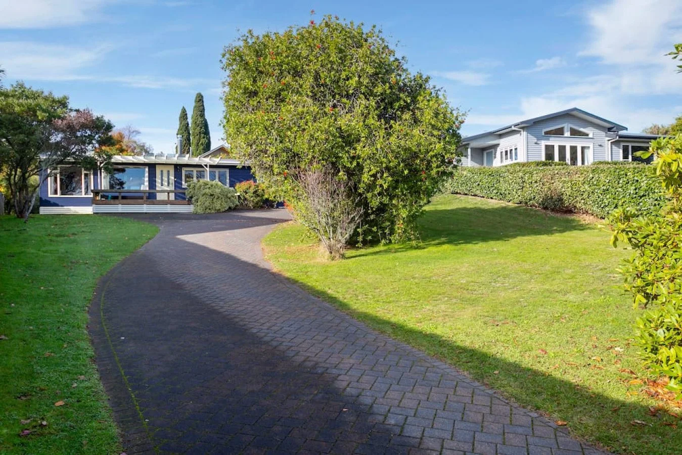 A curved driveway leads to two houses surrounded by green grass, trees, and bushes, under a partly cloudy sky.