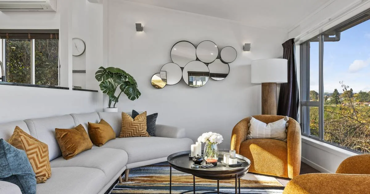 Modern living room with beige sofa, mustard and blue accent pillows, a gold armchair, a large window with a view, a round black coffee table with candles and flowers, a potted plant, and circular mirror decor on the white wall.