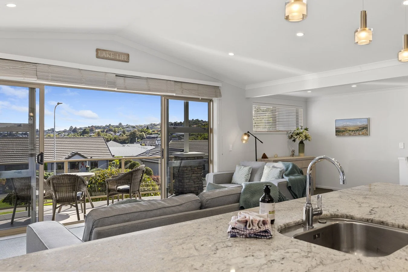 Living room with sliding glass door leading to a balcony, overlooking a neighborhood and hills in the distance. The interior features white walls, a gray sofa with pillows, a wooden sideboard with a potted plant, a modern lamp, and a mounted painting
