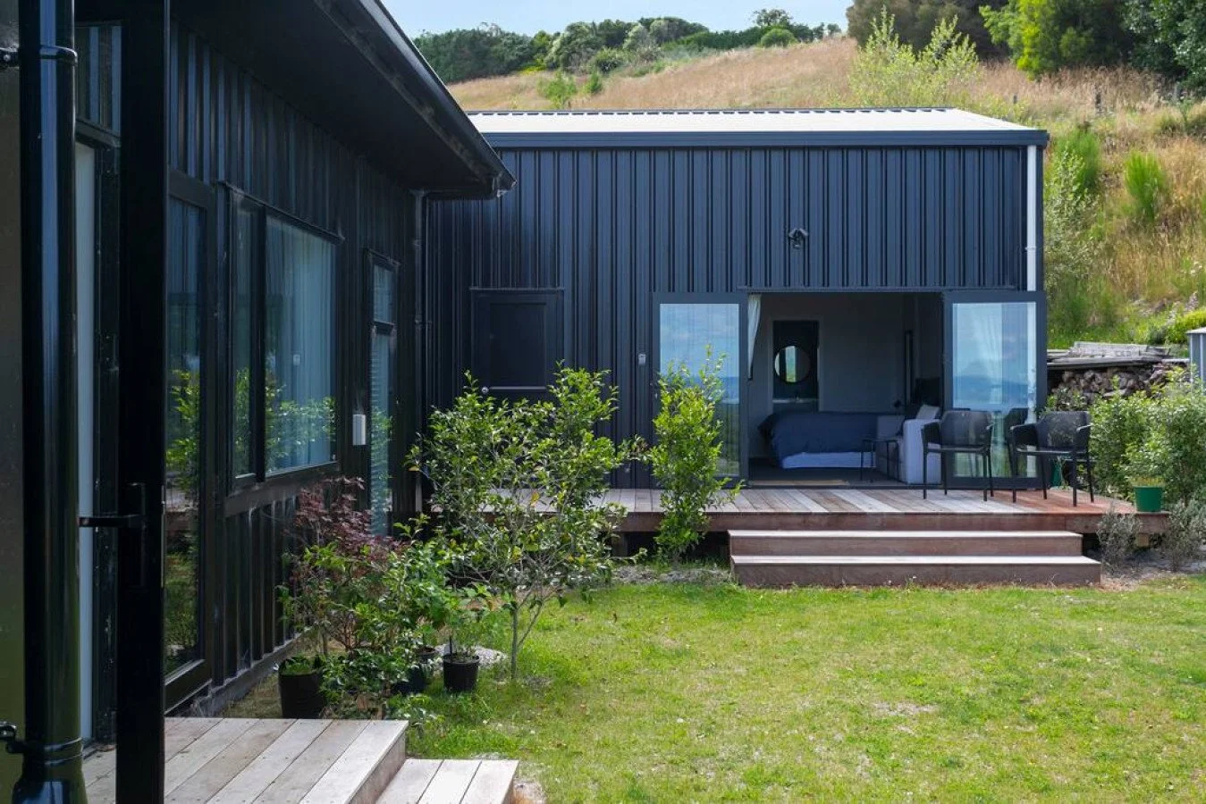 Modern black metal house with a small elevated wooden deck, open sliding glass doors, and outdoor seating, surrounded by green grass and trees in the background.