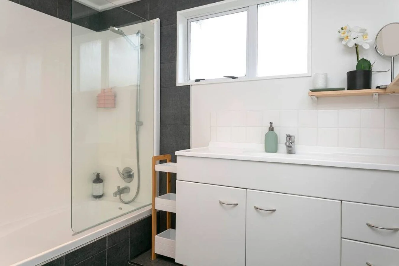 Bathroom with bathtub, glass shower screen, white vanity with storage drawers, window, and shelf with decorative items including a potted orchid, mirror, and containers.