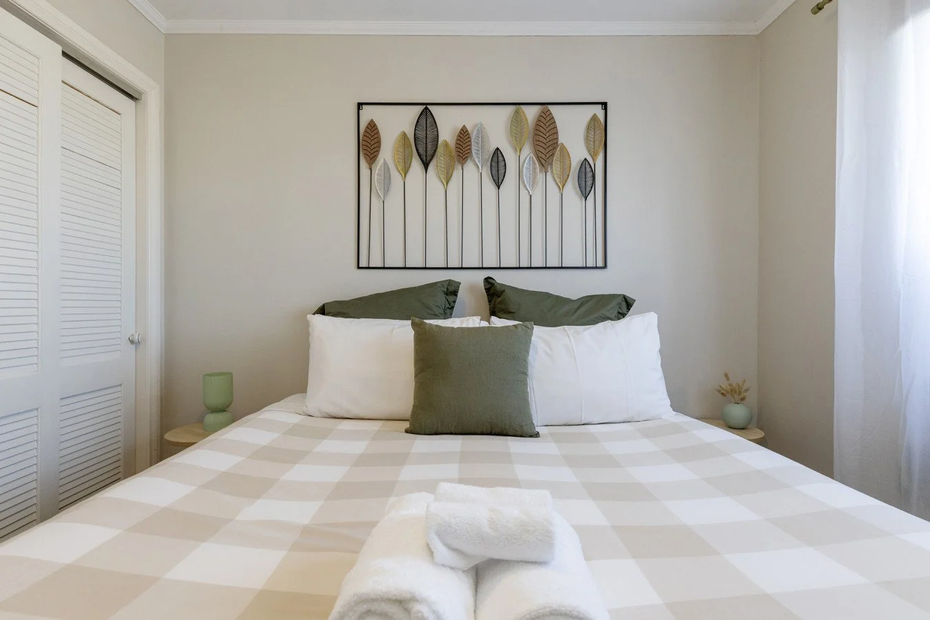 Bedroom with a bed that has folded towels on it, decorative pillows, and wall art of leaves above the bed. Side tables with vases and a closet with louvered doors.
