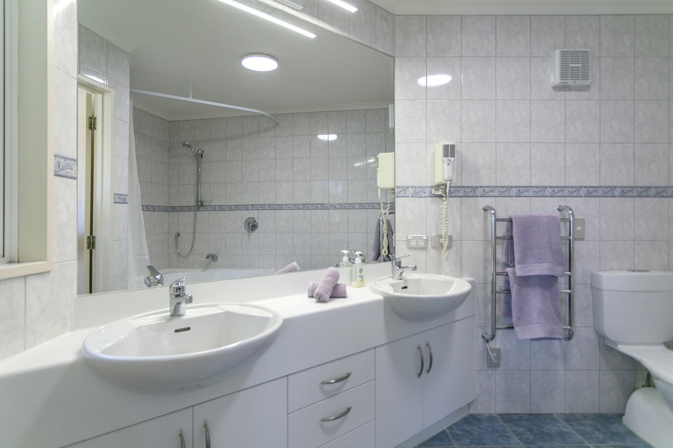 A clean, modern bathroom featuring two white sinks with silver faucets, a large mirror, a towel rack with purple towels, a wall-mounted hairdryer, a toilet, and a shower area with a showerhead and a curtain, all with tiled walls and floor.