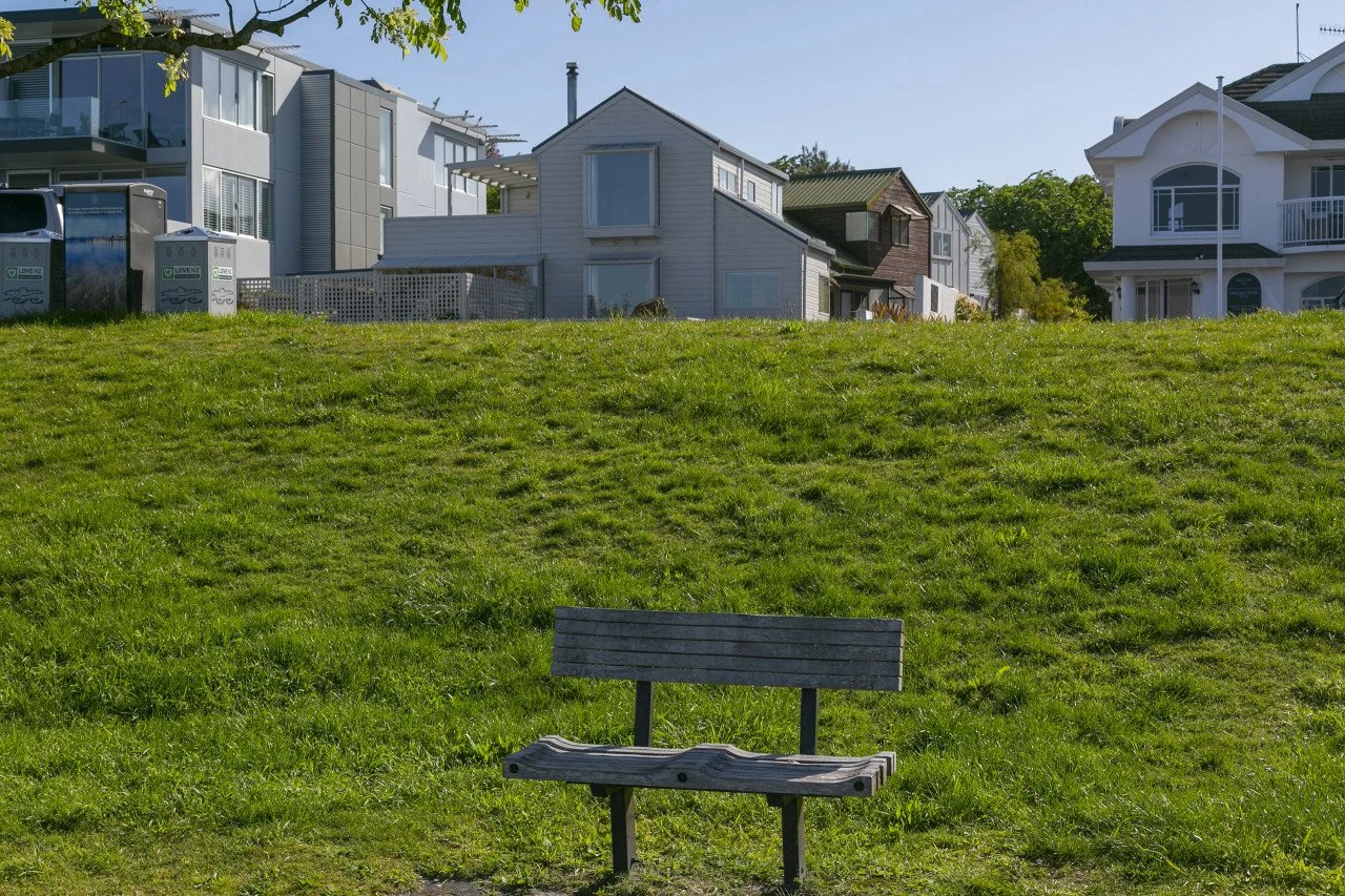 Empty wooden park bench in a grassy area with houses and buildings in the background under a clear blue sky.