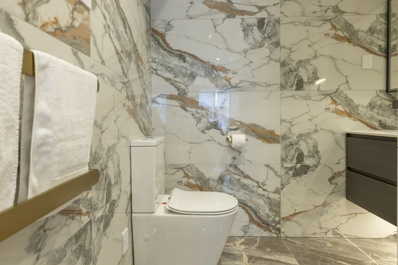 Modern bathroom with marble walls and floor, toilet with closed lid, towel rack with white towels, and a dark gray vanity with drawers.
