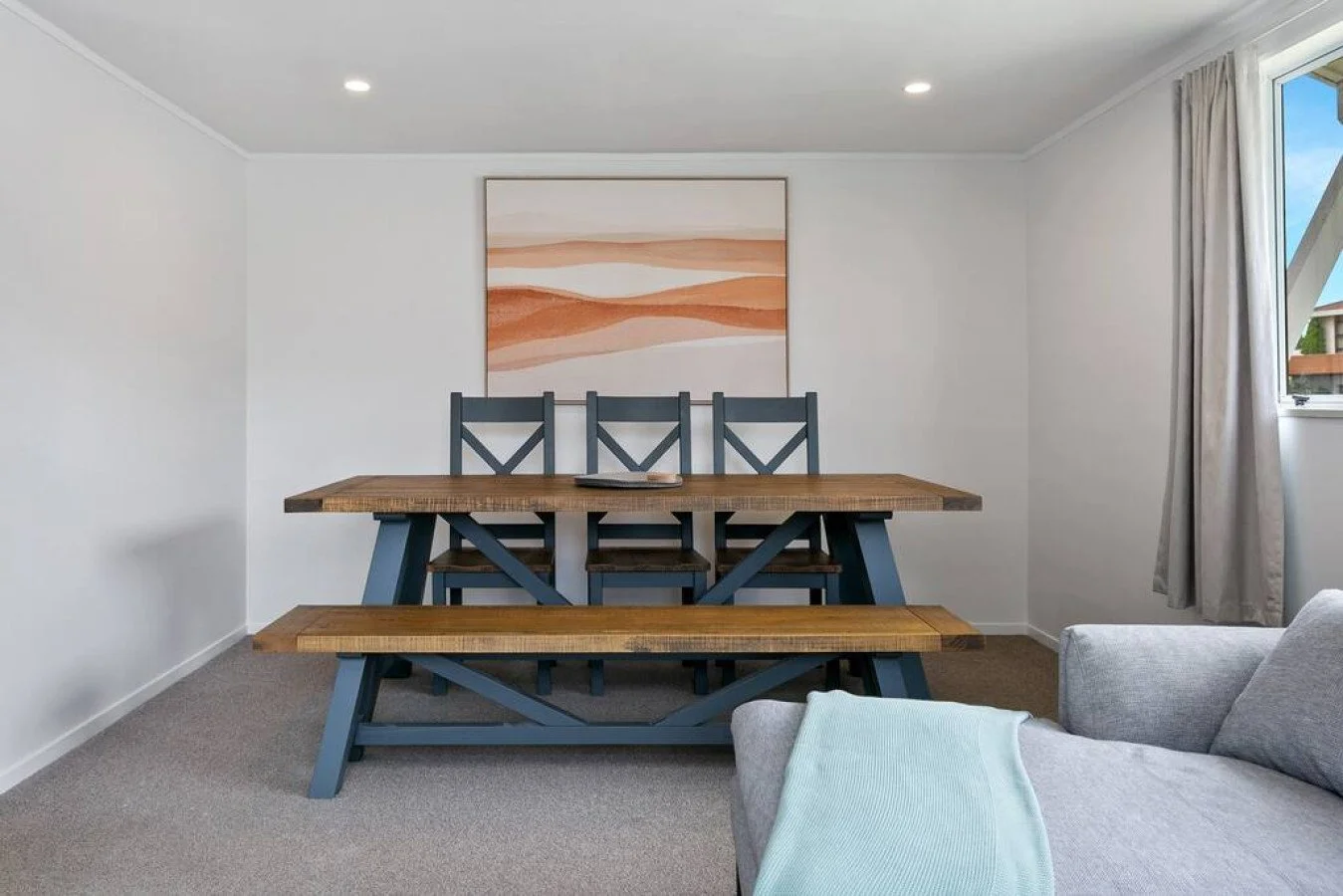 Dining room with a wooden table, blue chairs, and a bench, artwork on the wall, window with curtains, and part of a gray couch with a light blue blanket.