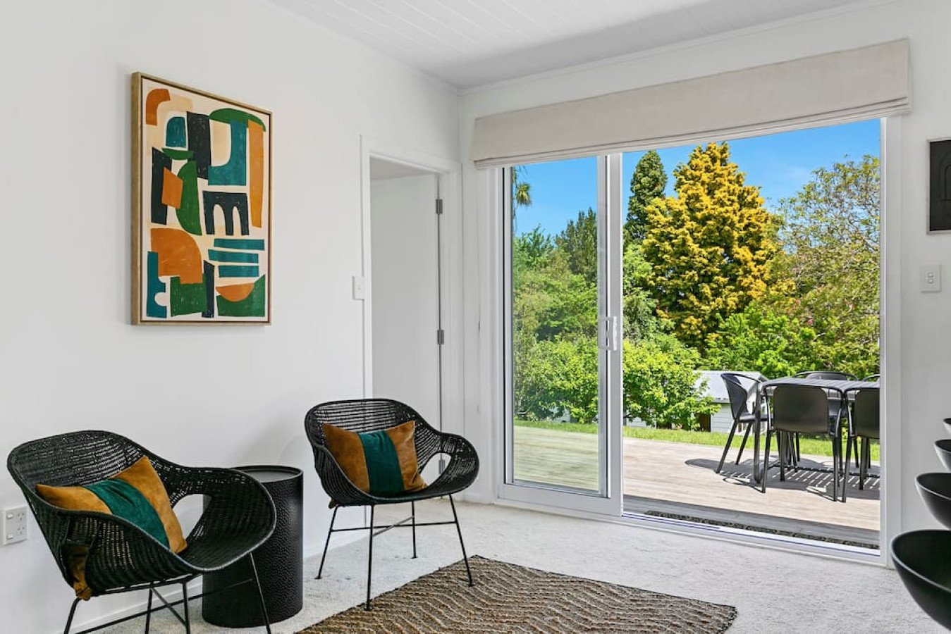 A bright living room with white walls and carpet, black wicker chairs with colorful cushions, abstract artwork on the wall, and large sliding glass doors leading to an outdoor patio with a table and chairs, overlooking green trees and blue sky.