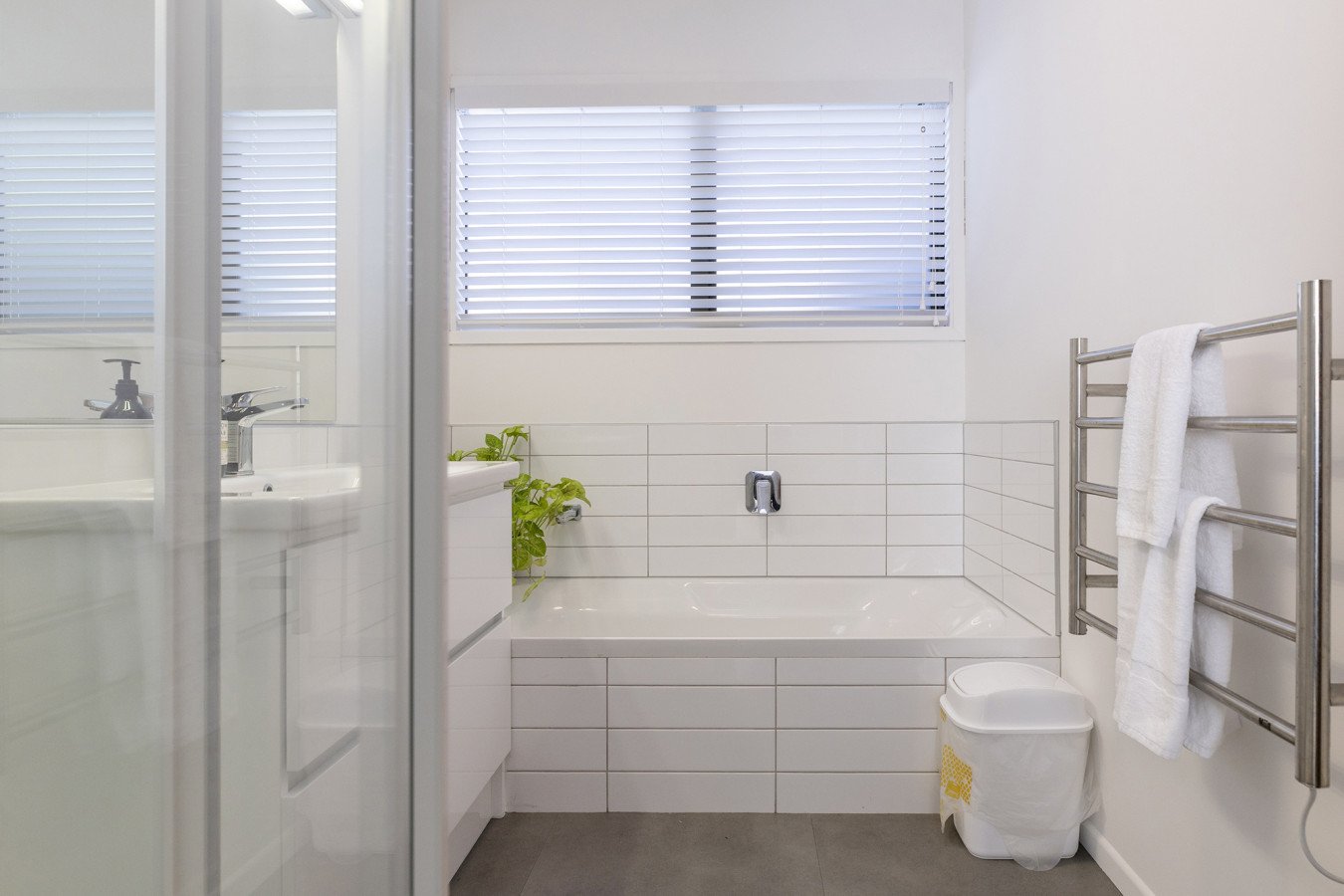 Modern bathroom with white tiles, a bathtub, a window with blinds, a towel rack with towels, a trash bin, a plant, and a shower door.