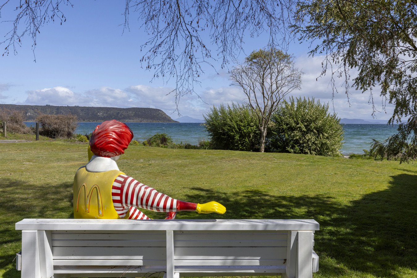 A outdoor scene with a McDonald's Ronald McDonald statue sitting on a white bench, facing a lake with hills in the distance. The sky is partly cloudy and there are trees and shrubs around.