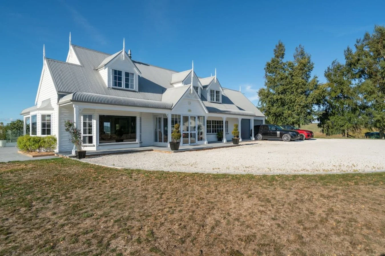 A large white house with a distinctive pointed roof design, front porch, and multiple large windows. There are a few cars parked outside on a gravel driveway, with large trees in the background and a clear blue sky.