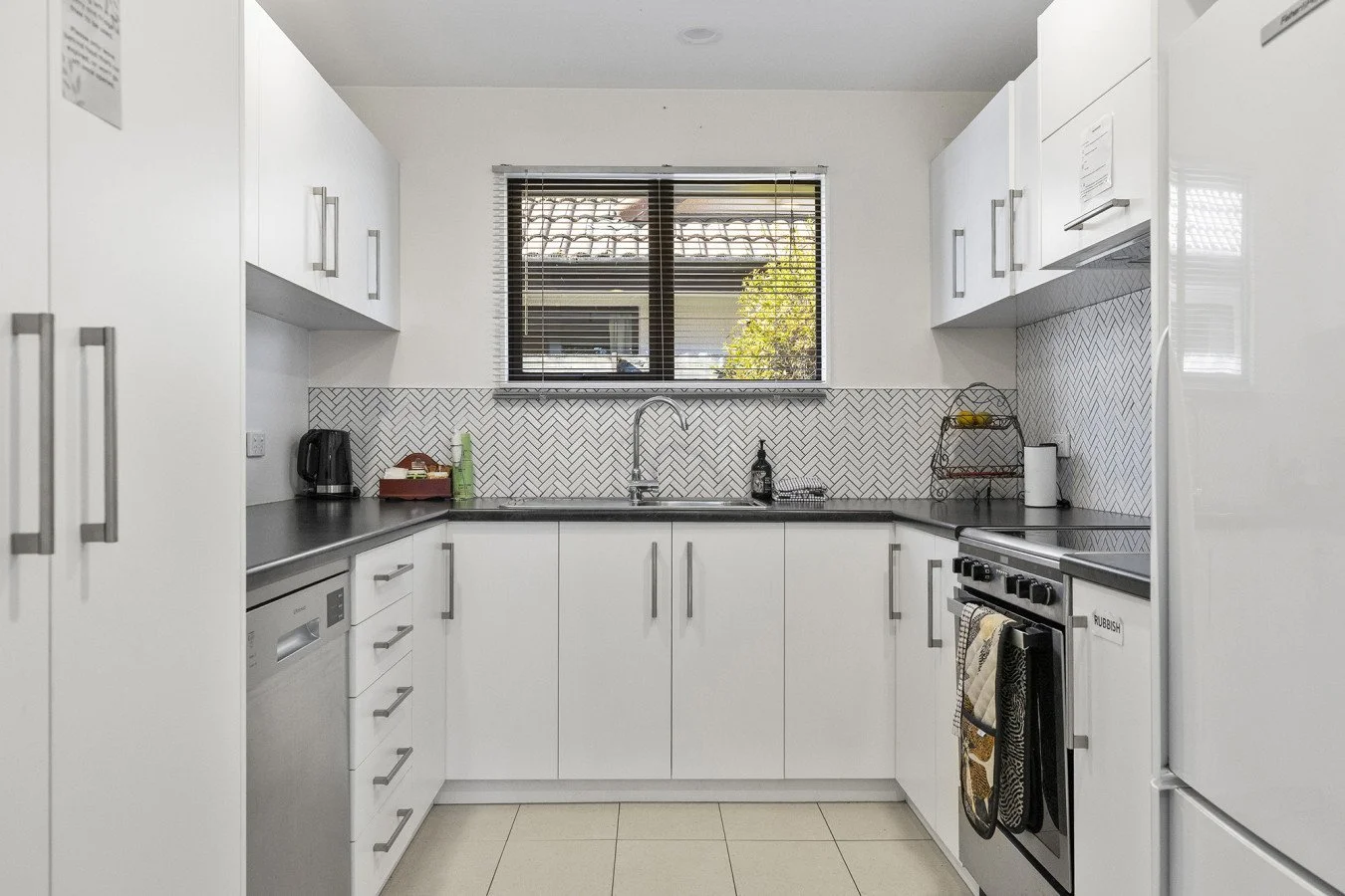 A clean kitchen with white cabinets, a black countertop, a window above the sink, a black kettle, a dish rack, an oven, a dishwasher, and a fridge.