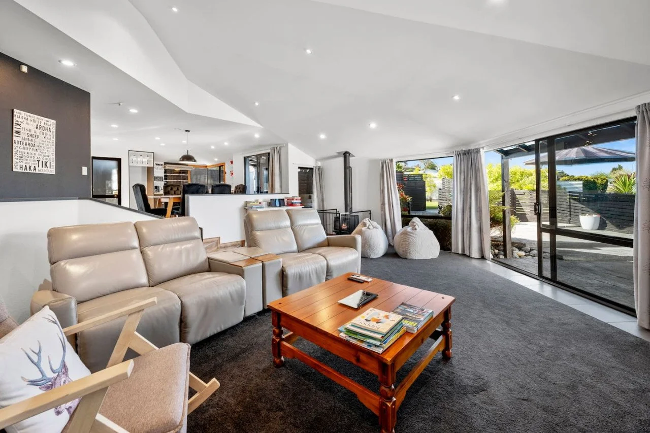 Living room with beige leather sofas, a wooden coffee table with magazines, a gray carpet, and large sliding glass doors leading to an outdoor patio with greenery.