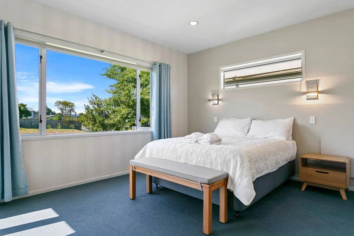 Bright bedroom with large window showing trees and blue sky, queen bed with white bedding and pillows, small wooden nightstand, bench at foot of bed, and wall-mounted lights.
