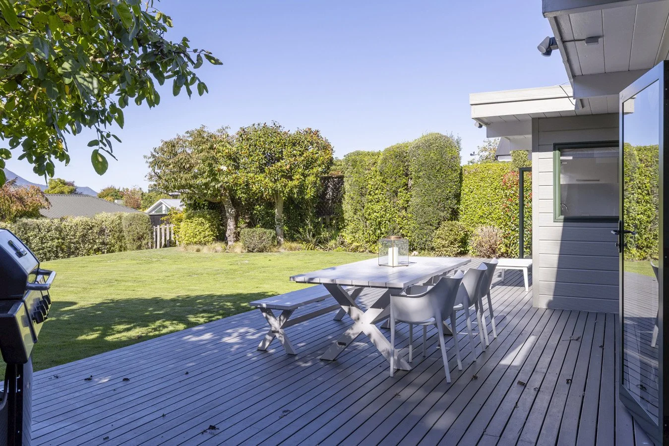 View of a backyard patio with a dining table and chairs, surrounded by greenery and trees, with a lawn and a backyard fence in the background.