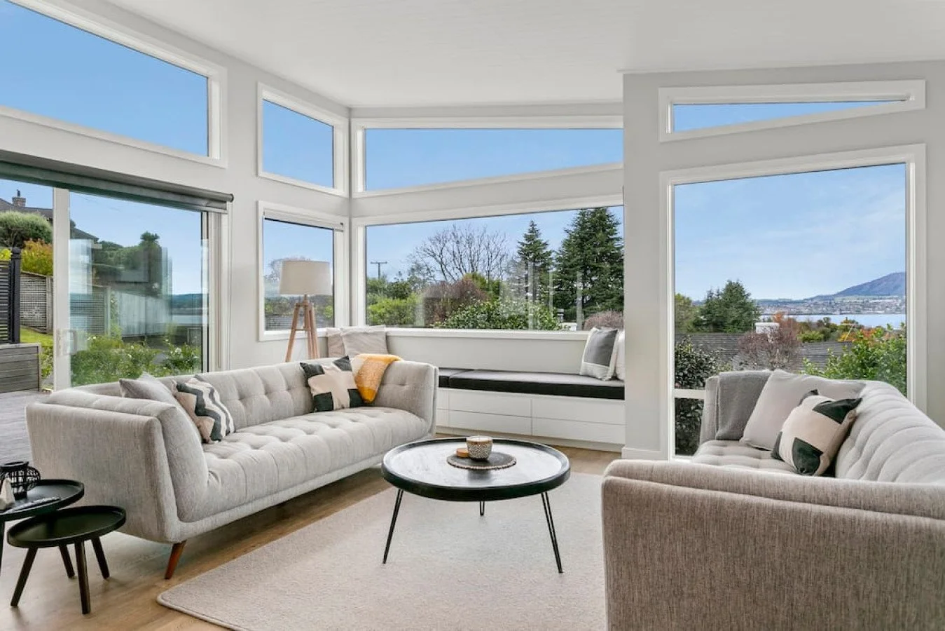 Bright living room with large windows showcasing trees, mountains, and blue sky, furnished with beige sofas, a black round coffee table, and a window seat with cushions.